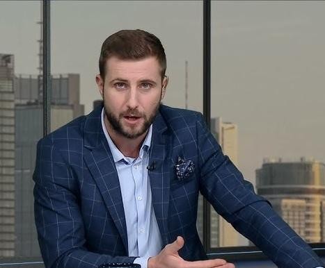 A professional, sharp, and engaging portrait of Miłosz Kłeczek, a Polish journalist and reporter. He should be depicted in a studio setting, possibly with subtle background elements hinting at a newsroom or television studio (e.g., blurred monitors, subtle lighting). The image should convey professionalism and confidence, reflecting his role as a public figure in media. He should be looking directly at the viewer with a serious but approachable expression. The style should be realistic, high-resolution, and suitable for a biographical article. Avoid any exaggerated features or caricatures.