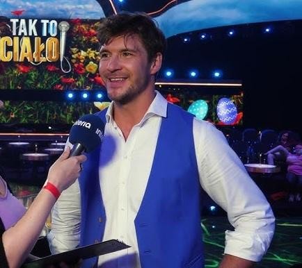 Mikołaj Roznerski na planie filmowym, w trakcie zdjęć lub rozmawiający z ekipą, w dynamicznej pozie, oddającej energię jego pracy.