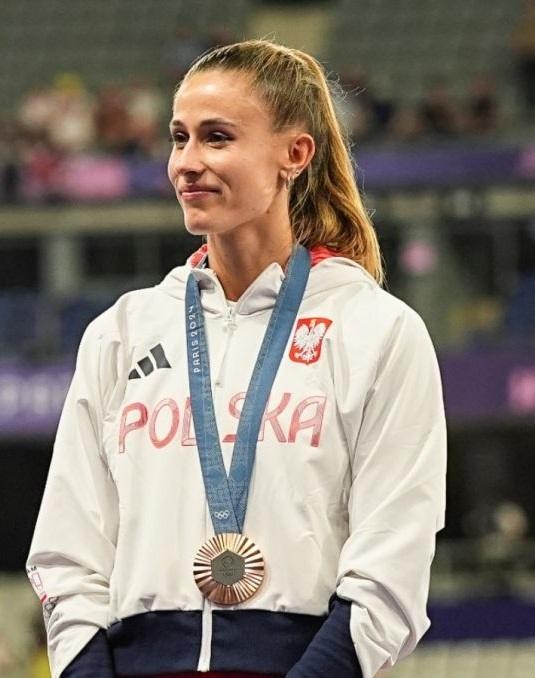 Natalia Bukowiecka (Kaczmarek) celebrująca zwycięstwo na bieżni, z szerokim uśmiechem i uniesionymi rękami, na tle rozmytego stadionu.