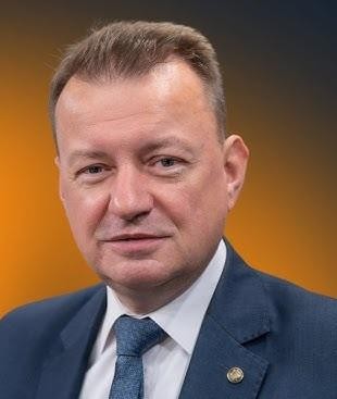 Mariusz Błaszczak jako Minister Obrony Narodowej, w towarzystwie żołnierzy, oglądający sprzęt wojskowy na poligonie.