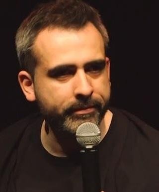 Abelard Giza podczas występu stand-upowego, z ekspresyjną mimiką i gestykulacją, na scenie oświetlonej reflektorami.