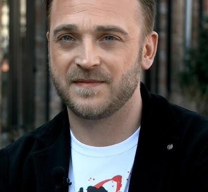 Mateusz Gessler na planie programu kulinarnego, w otoczeniu uczestników lub innych jurorów, aktywnie gestykulujący.