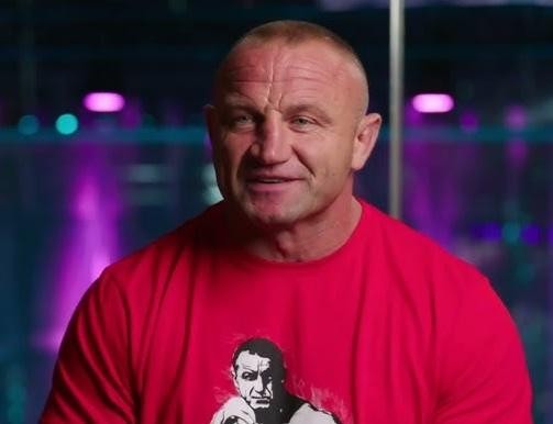 Mariusz Pudzianowski podczas walki MMA w oktagonie, w dynamicznej akcji z widocznym przeciwnikiem, na tle rozmytego tłumu i świateł areny.
