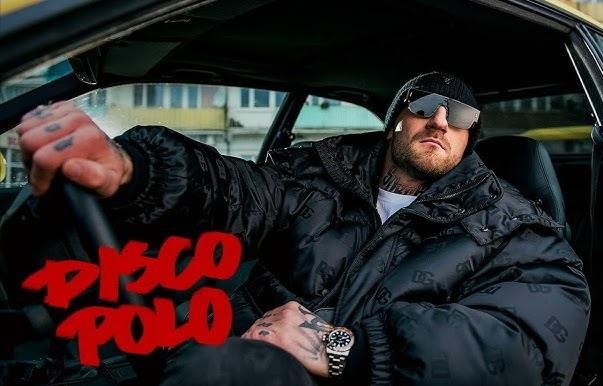 Kizo (Patryk Woziński) w dynamicznej pozie na scenie podczas koncertu, trzymający mikrofon, oświetlony kolorowymi światłami, symbolizujący jego sukcesy w karierze muzycznej.