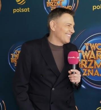 Krzysztof Ibisz w studiu telewizyjnym, energicznie prowadzący program, z uśmiechem i światłami w tle.
